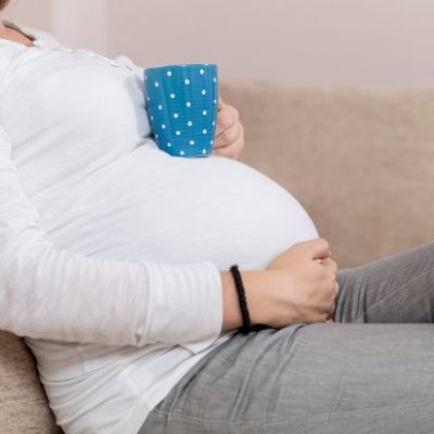 A woman in a white top reclines on a couch, resting a blue coffee cup on her pregnant stomach